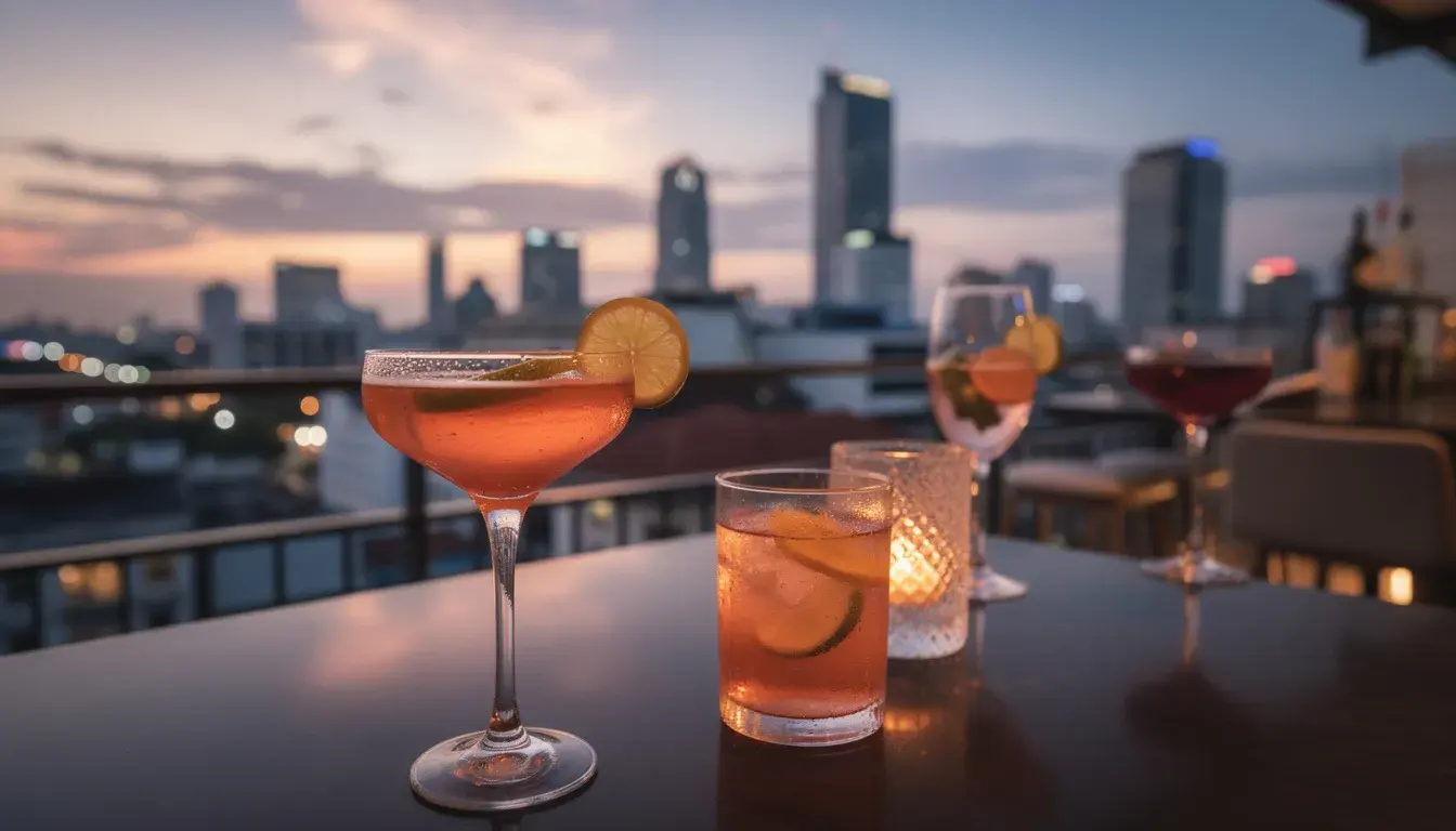 The image depicts a vibrant rooftop bar scene overlooking the Bangkok skyline at dusk, with city lights starting to twinkle against a colorful sky. In the foreground, cocktail glasses are elegantly placed, inviting visitors to enjoy the lively nightlife and stunning views of Thailand's capital.